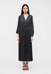 Μακρύ μαύρο πλισέ φόρεμα με βαθύ V-ντεκολτέ, μακριά μανίκια και υφή με λαμπερή όψη. Φορεμένο με μαύρα σανδάλια με λουριά.