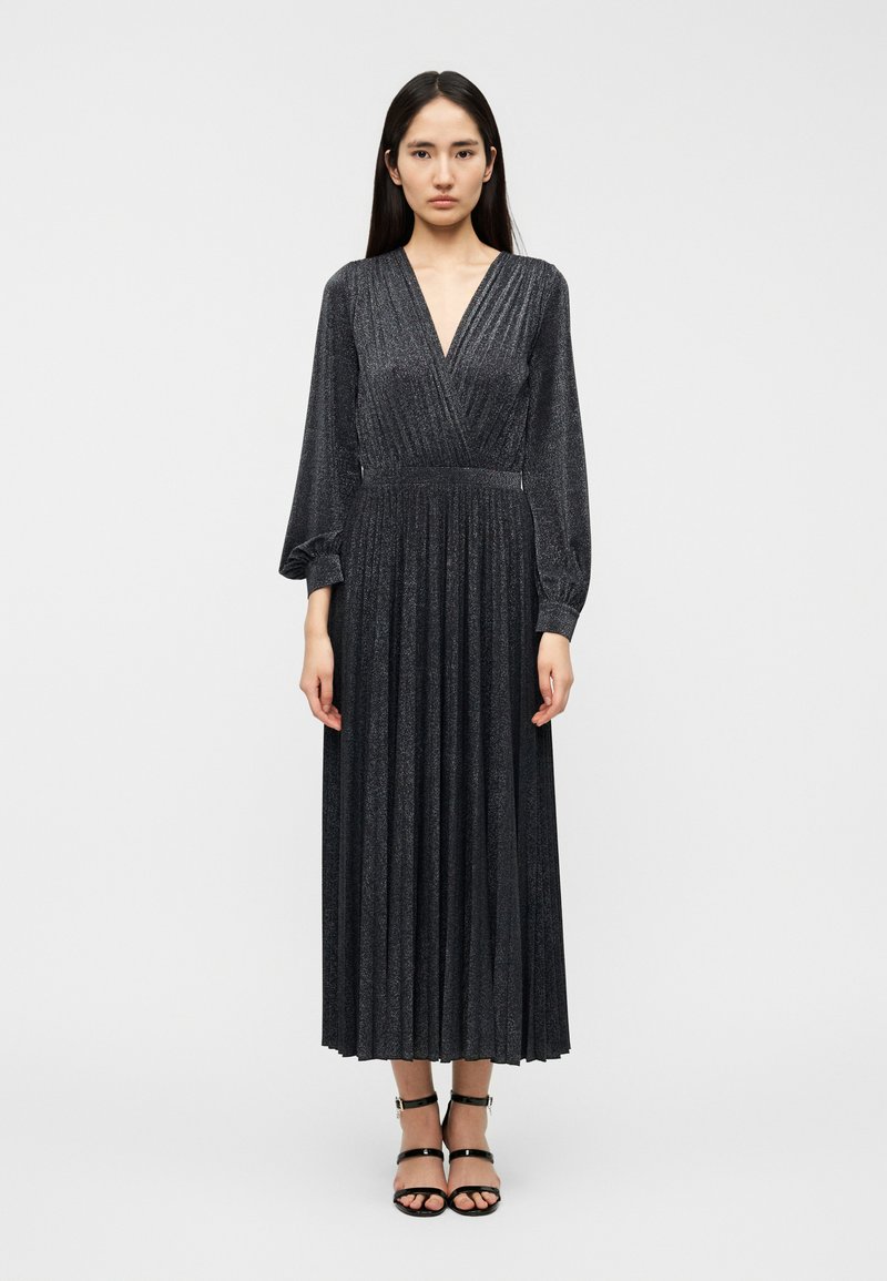 Μακρύ μαύρο πλισέ φόρεμα με βαθύ V-ντεκολτέ, μακριά μανίκια και υφή με λαμπερή όψη. Φορεμένο με μαύρα σανδάλια με λουριά.