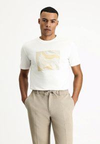 Camiseta blanca de manga corta con un diseño cuadrado abstracto en oro y blanco. Combinada con pantalones beige estampados y cintura con cordón.