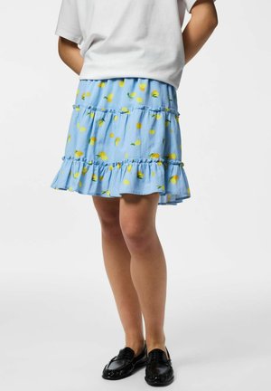 Biely top v kombinácii so svetlomodrou sukňou s citronovými vzormi a volánmi, nosený s čiernymi loafers na stojacej osobe.