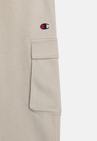 Lys beige sweatshirt med en myk tekstur, som har en sidelomme og en liten brodert logo i rødt, svart og hvitt på ermet.