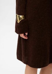 Palton din lână maro, pufos, cu un accent metalic auriu pe manșetă. Mâneca este ușor largă, lăsând să se vadă o parte dintr-o mână.