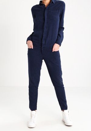 Person standing with hands in pockets wearing a navy blue button-up shirt, matching pants, and white high-top sneakers.