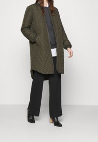 Persona che indossa un lungo giubbotto imbottito verde oliva, pantaloni neri a gamba larga, stivali neri con tacco e maglie sovrapposte grigie e bianche.