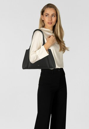 Borsa a mano in pelle nera con una forma strutturata, dotata di due manici lunghi e un logo discreto. Indossata casualmente sopra una maglia chiara.