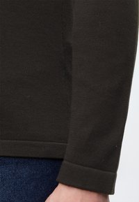 Gros plan sur le poignet et le bas d'une manche d'un vêtement en maille à manches longues de couleur foncée porté sur un jean en denim bleu.
