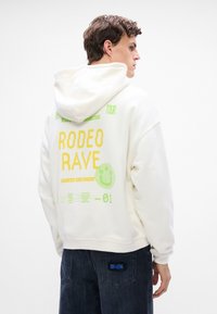 Balta kapuce ar zaļiem un dzelteniem grafiskiem attēliem, kurā ir teksts "RODEO RAVE" un smaida ikona. Mīksta tekstūra, brīva piegriezuma, kapuce, saskaņota ar tumšām džinsām.