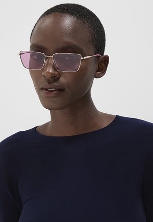 Persona con i capelli corti che indossa occhiali da sole rettangolari con lenti viola e una maglietta blu scuro su uno sfondo chiaro semplice.