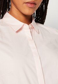 Gros plan d'une personne aux cheveux tressés portant une chemise rose pâle boutonnée avec un col classique, montrant le bas du visage et le cou.