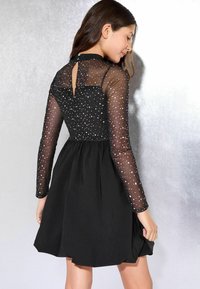 Schwarzes Kleid mit langen, transparenten Ärmeln, die mit silbernen Punkten verziert sind. Der Rücken verfügt über eine Schlüssellochöffnung und einen Knopfverschluss. Ausgestellter Rock.