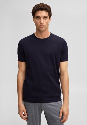 Giovane uomo con capelli ricci e scuri che indossa una camicia aderente blu navy a maniche corte e pantaloni grigi, in piedi davanti a uno sfondo grigio uniforme.