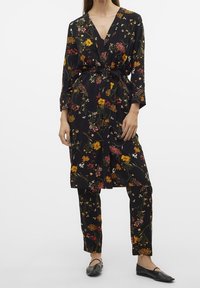 Svart blommig jumpsuit med djup V-ringning, långa ärmar, knytbart midjeband och matchande byxor med olika färgglada blommönster.