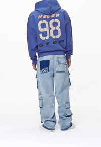Sudadera morada con gráfico "NEVER STOP 98"; jeans cargo de color azul claro con detalles de parches; corte relajado; puños acanalados; zapatillas blancas.