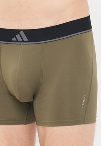 Masculine boxer šortki v oljčno zeleni barvi, izdelani iz gladkega materiala, z črnim pasom, okrašenim s sivo barvo ter logotipom "AEROREADY" na strani.