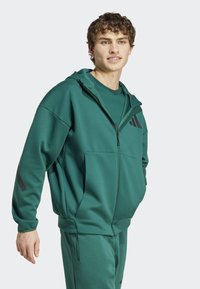 Zeleni zip-up atletski pulover s visokim ovratnikom, sprednjimi žepi in črnimi oglatimi akcenti. Usklajen s hlačami; gladka tekstura.