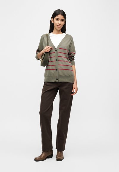Πλεκτή ζακέτα σε ελιά πράσινο με κόκκινες ρίγες, κούμπωμα με κουμπιά και μακριά μανίκια, συνδυασμένη με λευκό T-shirt, καφέ παντελόνι και καφέ μποτάκια μέχρι τον αστράγαλο.
