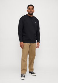 Svart sweatshirt med rundad halsringning, med en liten logga på bröstet. Kombinerad med khaki-byxor och svarta sneakers med vita sulor.