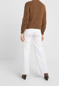 Bruine gebreide trui met kabelgebreide mouwen, gecombineerd met witte wijde broek en chunky hakken. Minimalistische achtergrond.