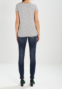 Camiseta gris de manga corta con un sutil estampado blanco, combinada con jeans oscuros ajustados y zapatos de tacón verdes. Vista trasera, fondo neutro.