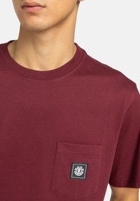 Camiseta de algodón burdeos con cuello redondo, mangas cortas y un bolsillo frontal que presenta un logo de etiqueta negra. Tela suave y sin textura.