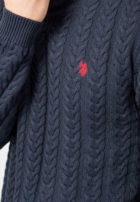 Mörkblå kabelstickad tröja med ett upphöjt mönster och en liten röd logotyp på framsidan. Ribbad mudd och krage fulländar designen.