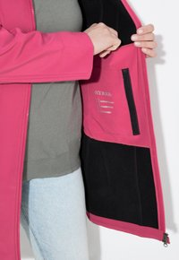Růžová vnější bunda rozepnutá, aby odhalila šedou podšívku a černé akcenty. Mezi funkce patří zipová kapsa a štítky pro vodu, vítr a prodyšnost.