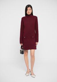 Maroon sweater dress with ribbed sleeves, two front pockets, and gold buttons. Paired with white low-heeled shoes and a small black handbag.