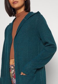 Teal huva cardigan med en texturerad stickning, öppen framsida och två framsidans fickor. Designen har långa ärmar och avslappnad passform.