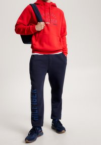 Felpa rossa con stampa grafica in navy, abbinata a pantaloni della tuta navy con un logo lungo la gamba e scarpe da ginnastica blu.