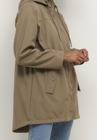 Impermeabile verde oliva con cappuccio, bottoni a pressione e tasche laterali. Realizzato in tessuto liscio e impermeabile. Presenta una vestibilità ampia e rilassata.