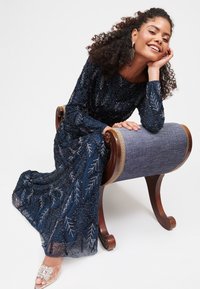 Marineblaues Kleid mit verzierten silbernen Blättern, langen Ärmeln, fließendem Stoff und geschichteter Textur. Getragen mit einer Sandale mit transparentem Absatz.