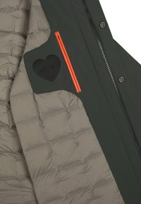 Giacca a vento verde e beige con texture trapuntata, che presenta una tasca con zip arancione e un accento nero a forma di cuore sul lato.