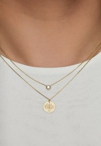 Des colliers en plaqué or avec un pendentif circulaire arborant un motif d'arbre et un petit cristal clair sur une chaîne délicate, portés sur un haut blanc.