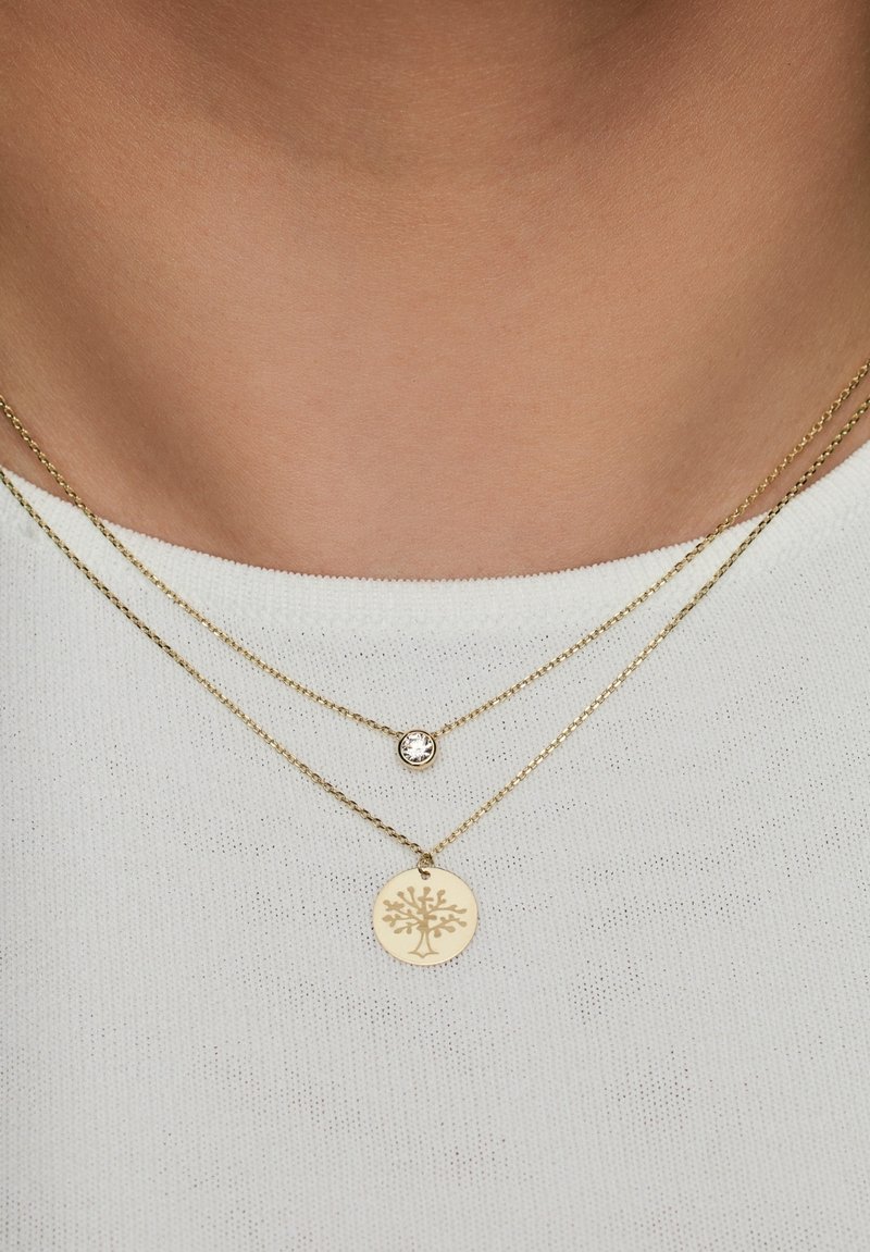 Des colliers en plaqué or avec un pendentif circulaire arborant un motif d'arbre et un petit cristal clair sur une chaîne délicate, portés sur un haut blanc.