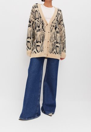 Person wearing beige and black snake-patterned cardigan, white top, wide-leg blue jeans, and pointed shoes on white background.