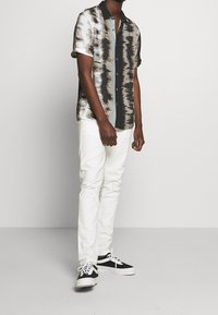 Košile s krátkým rukávem na knoflíky s texturovaným, abstraktním vzorem v černé, bílé a světle modré, doplněná bílými kalhotami a černými teniskami.