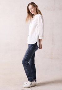 Camicia bianca in cotone con maniche arrotolate, abbinata a jeans in denim blu scuro e sneakers bianche con accenti grigi.