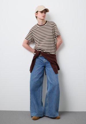 Persona joven con gorra beige, camiseta de rayas marrón y beige, pantalones vaqueros anchos azules y suéter marrón atado a la cintura, de pie contra una pared blanca.