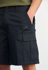 Black cargo shorts made from textured fabric with side pockets and a visible Nike logo in matching dark green, featuring a button closure.