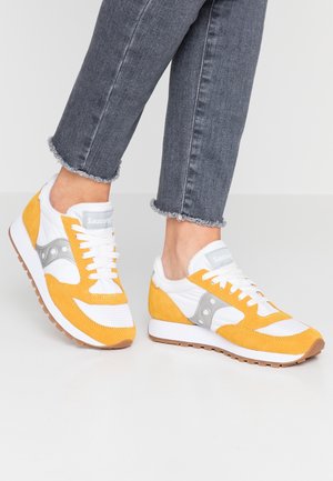 Voeten met gele, witte en grijze Saucony sneakers met witte veters, gecombineerd met grijze versleten enkellange spijkerbroek op een witte vloer.