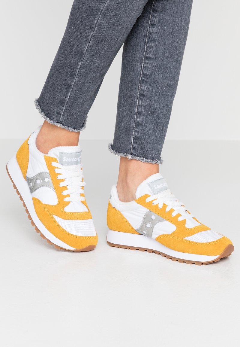 Pieds chaussés de baskets Saucony jaunes, blanches et grises avec des lacets blancs, associées à un jean gris effiloché à la cheville, sur un sol blanc.