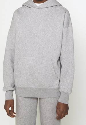 Personne portant un sweat à capuche gris clair avec poche avant et un pantalon de jogging gris assorti sur un fond uni.