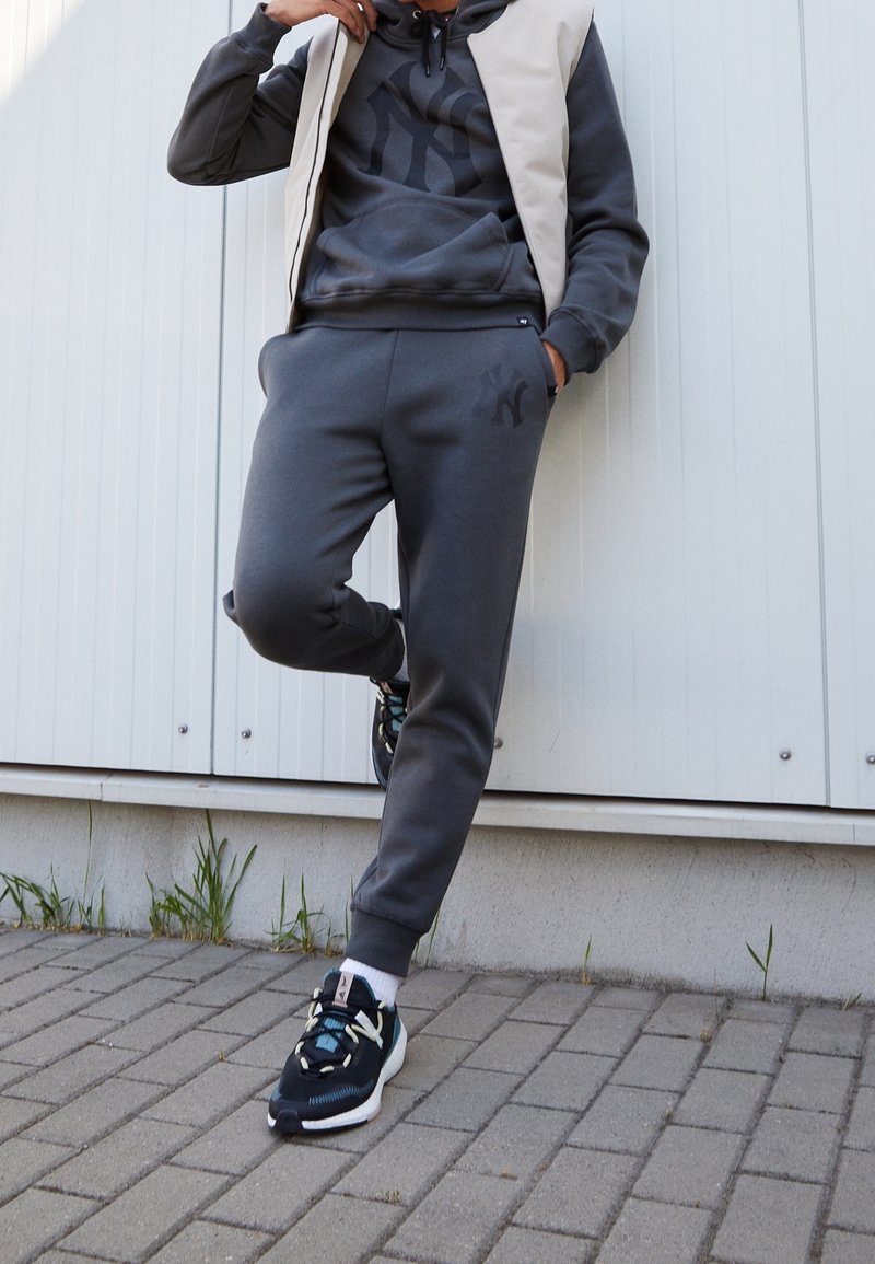Sweat-shirt gris avec un logo, pantalon de jogging gris et chaussures de sport noires avec des accents turquoise. Un gilet beige recouvre la tenue.