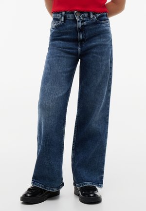 Person trägt dunkelblaue, weit geschnittene Jeans, rotes Oberteil und schwarze klobige Schuhe und steht vor weißem Hintergrund.