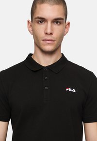 Svart pikétröja med krage, med tre knappslå, texturerad tyg och en liten broderad FILA-logotyp i röd och vit.