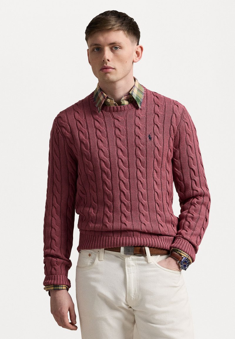 Crimson cable-knit sweater with ribbed cuffs and hem, worn over a plaid collared shirt; paired with white pants and a metal watch.