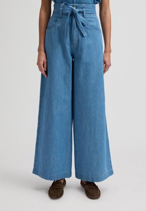 Femme portant un pantalon en jean bleu taille haute, à jambes larges, avec une ceinture nouée à la taille et des mocassins marron, les mains reposant le long du corps.
