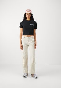 Svart kort t-shirt med "PEQUIS" tryck, beige cargobyxor med sidofickor och flerfärgad bucket hat, tillsammans med silvriga sportskor.