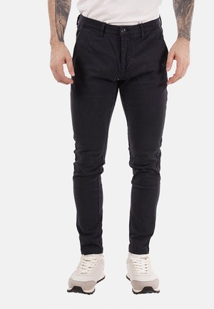 Uomo che indossa pantaloni neri slim fit, sneakers bianche e una camicia bianca, in piedi con le mani rilassate lungo i fianchi.