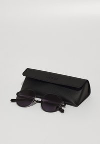 Ochelari de soare rotunzi negri cu lentile închise, accente metalice și o carcasă din piele cu o suprafață texturată și logo-ul „GUESS” embossat.